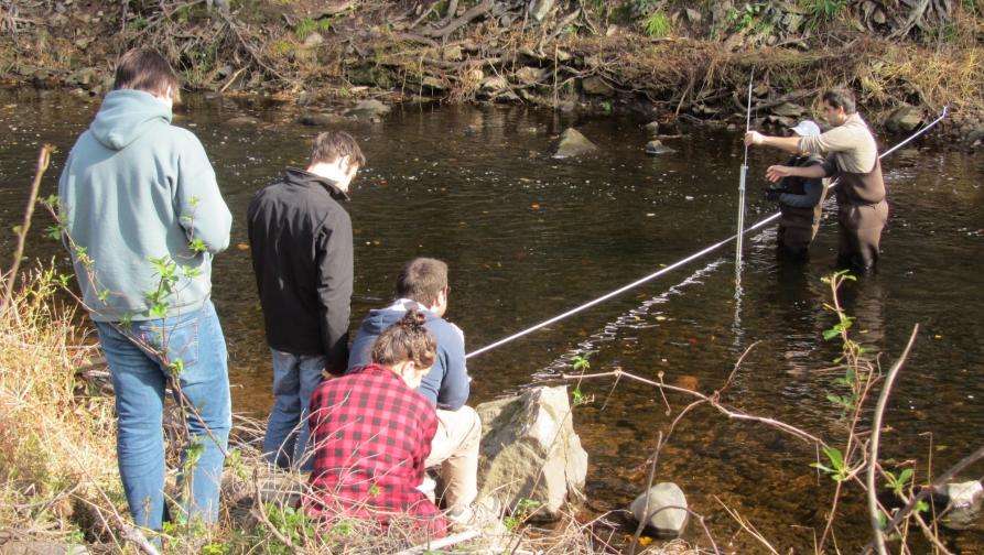 Earth science students sitting by a creek and watching a demonstration of someone setting up a contraption. 